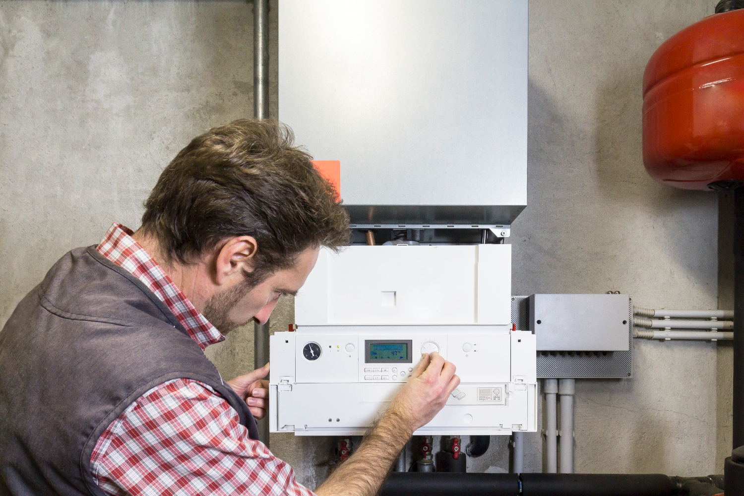 A close-up, eye-level indoor shot shows a male technician in a red and white checkered shirt and a dark gray vest, leaning in and working on a white, wall-mounted boiler system. He is using both hands to adjust a dial on the control panel, which has a small blue digital display. In the background, there are gray concrete walls, a large red tank, and various pipes and electrical conduits.
